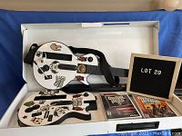 Image of two white Guitar Hero guitar controllers adorned with colorful rock-themed stickers, laid inside a box with two Wii Guitar Hero game cases beside them