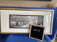 Frontal view of the professionally framed black and white photograph titled "Laneway Walk in the Fall" with a gold-color frame, displayed on blue fabric with a lot number sign.