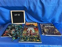 Full view of Warhammer 40,000 books spread on blue cloth surface with a group of painted miniature figurines on left side and Lot 53 sign board.