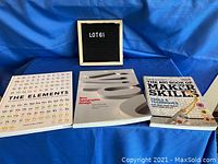 Three books arranged on blue cloth fabric: 'The Elements', 'Typographic Design', 'The Big Book of Maker Skills'. A black letter board with 'LOT 61' is displayed behind.