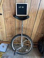 Front view of unicycle showing full 20-inch wheel, pedals, and black curved seat against wood panel background.