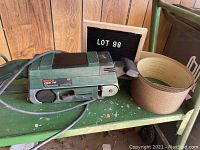 Bosch PBS 75 electric belt sander, green colored, placed on a green metal shelf with sanding belts next to it.