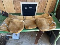 Front view of brown leather work belt with multiple suede pouches laid out on a table beneath a 'Lot 99' sign.