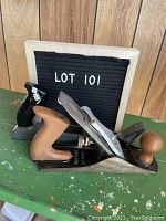 Two portable hand planers placed side by side on a green surface with a black board labeled LOT 101 in the background.
