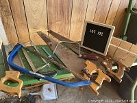 Photo showing multiple hand saws arranged on a wooden surface with a wooden miter box on the right side and a letter board marked LOT 102.