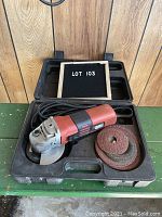 Black & Decker electric angle grinder in molded black plastic carrying case with abrasive discs inside, lot number 103 indicated on a small board.