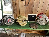 Photo showing three circular saws and one detached blade on a green workbench. Two Black & Decker saws on left, Skilsaw on right. Wall with vertical wood panels in background.