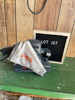 Front angle view of SKIL electric hand sander with attached dust filter bag, placed on green wooden surface with wood wall background, and Lot 107 board.