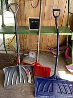 Photo of three snow shovels standing upright showing plastic lifters in gray, red, and blue colors with wooden and plastic handles.