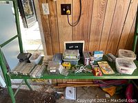 Photo showing various containers and boxes with assorted nails, screws, hooks, and hardware items arranged on a table with wooden panel background.