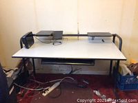 Front view of the white computer desk with black metal legs, elevated platforms on rear bar and a sliding keyboard tray underneath the main surface. Cord clutter seen under and around the desk.