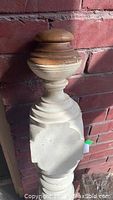 Close-up photo of the top portion of antique turned wood bannister post showing natural wood rounded finial and painted turned body against brick background.