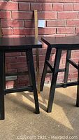 Two black wooden bar stools leaning against a red brick wall in sunlight, showing side and corner angles of the stools.