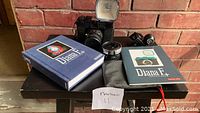 Photo showing the Diana F+ camera with flash, additional lens accessory, black carrying pouch, hardcover book and manual