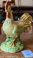 Front and side view of the hand-painted ceramic rooster showing detailed textured feathers and natural coloration including white, green, orange and red. It stands on a green base with sculpted grass detail.