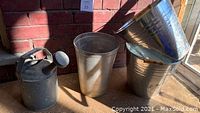 Photo showing two cylindrical metal buckets with wooden handles and one tall tin container with a metal handle, all vintage and metallic in appearance.
