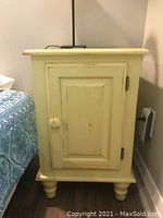 Front view of vintage hand made wood painted night table showing hinged door, wooden knob, turned legs, and distressed light cream paint.