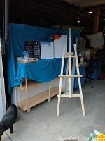 Photo showing wooden easel standing with canvas and art supplies on a table with blue cloth