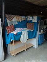 Wide shot showing the full display of linens and pillows arranged on stapled blue cloth draped over furniture. Includes multiple linens with lace edges and two decorative pillows in earthy colors and geometric designs.