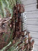 Stack of red clay bricks with two concrete decorative blocks placed on top, surrounded by ferns outdoors.