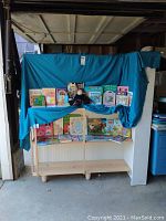 Wooden bookshelf with a blue cloth backdrop displaying a variety of children's books arranged on two shelves and a middle display shelf with a stuffed toy sitting on top.
