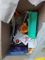 Top view of cardboard box packed with kitchen tools including orange coffee grinder, green salad spinner, plastic containers, and various small kitchen utensils.