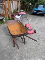Photo showing rusty metal wheelbarrow with green single tire, red and black cordless lawnmower behind it, and wire tomato plant stand nearby