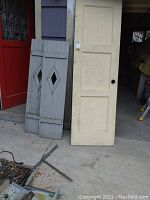 Five wooden doors and two wooden shutters leaning against building exterior, showing condition and paint colors.