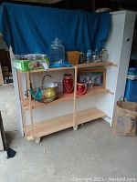 Photo showing shelving with multiple canning jars, two red outdoor candle holder jars, a large sun tea jar with a spout, a framed stained glass window art, and various decorative items.