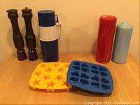 Pair of tall wooden salt and pepper mills, blue and white thermos, two tall cylindrical candles in red and light blue, and two silicone ice cube trays (one yellow with stars, one blue with assorted shapes) on wooden surface.