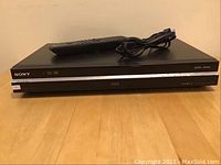 Front view of Sony DVD recorder with remote control and power cord placed on top of unit.