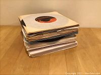 Stack of approximately 50 seven-inch 45 rpm vinyl records in paper sleeves on a wooden surface