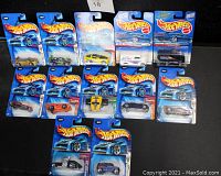 Photo showing 12 Hot Wheels cars in original blue packaging arranged on a black surface, featuring cars in yellow, orange, black, silver, and blue colors.