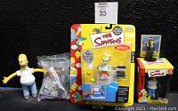 Photo showing five different Simpsons action figures and collectibles, with flexible Homer figure, mini Homer figure in packaging, Wendell figure in carded packaging, Tom Hanks figure with card, and Creepy Classic Homer figure in package.