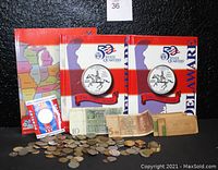 Wide view showing two 50 State Quarters coin books with Delaware on cover, assorted coins scattered in front, paper money, Olympic coin in packaging, coin wrappers and paperweight to right.