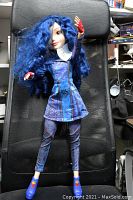Full view of the Evie Isle doll standing on a black mesh chair showcasing long blue curly hair, dressed in detailed blue-purple outfit with matching pants and red gloves and shoes.