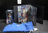 Binder open showing baseball cards sleeved and organized in clear sleeves, blue Dodgers swim trunks laid in front, and Rich Hill bobble-head in original box on left side.