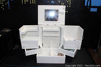 Open white wood jewelry box showing fold-up mirror, multiple compartments, and drawers with assorted jewelry inside.