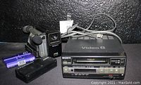 View of Sony Video 8 CCD-M8U camera recorder on top of Sony EV-C3 NTSC Video 8 cassette player, with battery charger adapter and sealed 60-minute video cassette tape beside them.