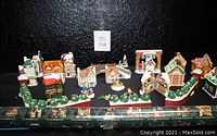 Front overall view of assorted ceramic small house village pieces and village bases with greenery, figurines, and a nativity piece, arranged in a linear display with rolled poster visible in front.