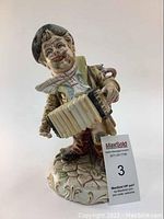 Front angled view of porcelain Capodimonte tramp figurine playing accordion on decorative base.