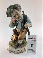 Full view front of Capodimonte porcelain hunter figurine with green hat, cup in hand, rifle on back, and original tag attached, on a decorated base.