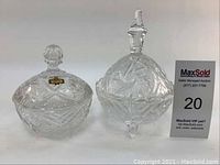 Two crystal candy dishes with lids on a white background, one taller with a pointed lid handle, the other smaller with a round knob.