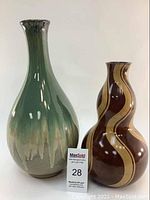 Two vases displayed side by side: a taller green ceramic vase and a shorter wooden vase with wavy stripe patterns, both shown upright on a neutral background.