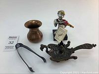 Photo showing a female figurine with an original tag, a brass vase, metal tongs, and an ornate cast metal decorative piece