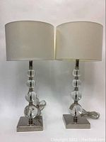 Front view of two identical ROXMO IKEA table lamps with white drum shades and chrome bases showing cords coiled below them.