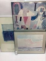 Photo of three framed poster prints, one with abstract geometric design with nature and architectural elements, one with soft pastel landscape, and one with abstract color blocks. Frames show visible wear.