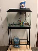 Full view of the 24-inch glass aquarium on a black metal stand with aquarium supplies inside including treatment bottles, fish net, and aquarium light on back wall.