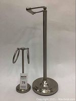 Two metal bathroom accessory stands, one tall toilet paper holder and one smaller double ring holder