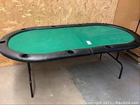 Poker table folded out and standing on metal legs, green felt playing surface visible with black padded edges and cup holders.